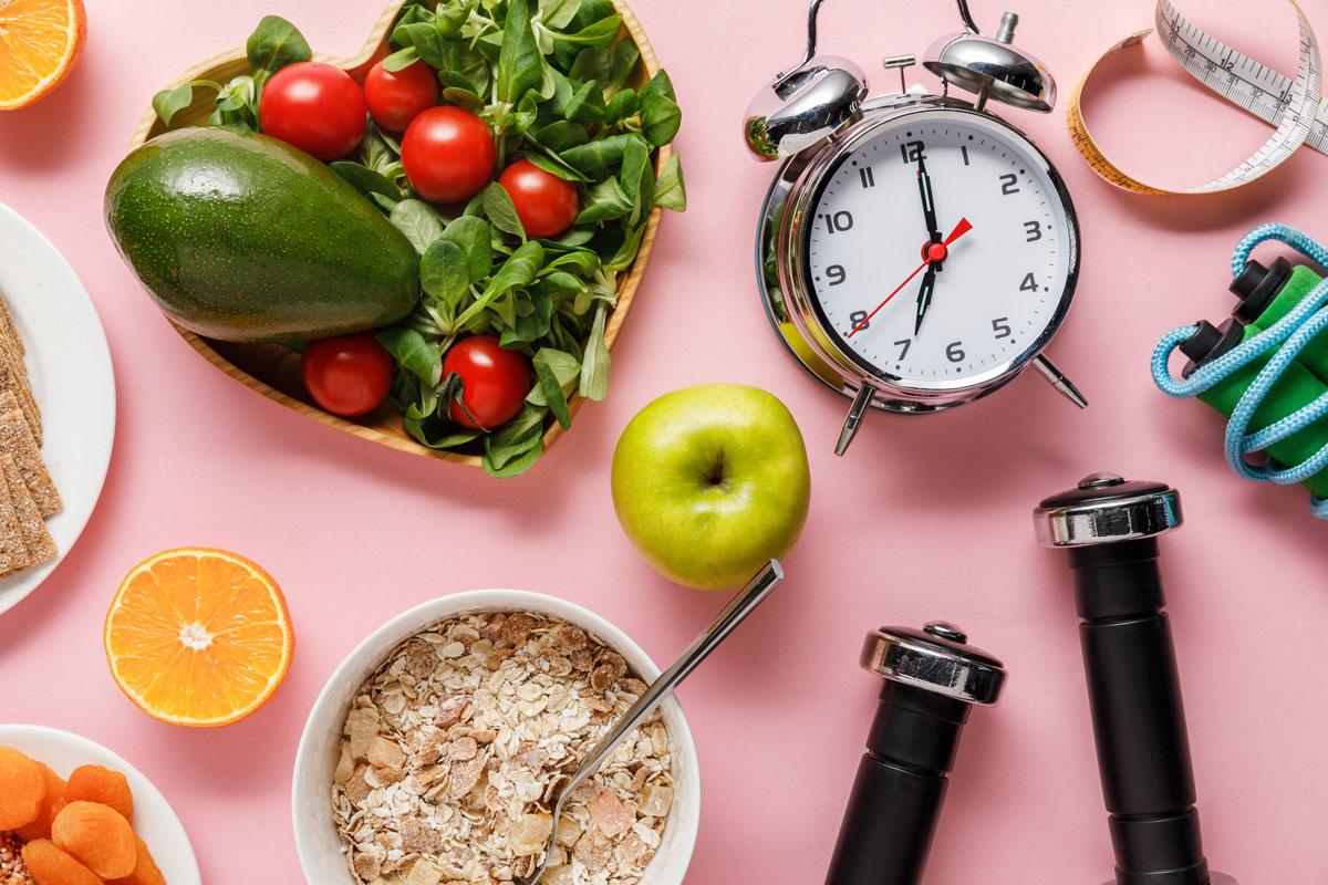 top view of fresh diet food, measuring tape, sport equipment and alarm clock on pink background