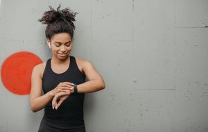 Jogging result. Happy african american woman with wireless headphones checks pulse on fitness tracker on gray wall background in city, copy space